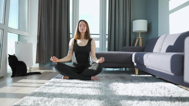 Young caucasian woman in black jumpsuit sitting in lotus position and meditating, yoga at home. Black cat sits on the background.