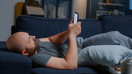 Upset man caused by break up lying on sofa at home browsing on smartphone. Lonely male feeling disappointed with heartbroken, suffering from mental problems, psychological trauma concept