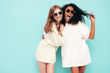 Two young beautiful smiling international hipster female in trendy summer clothes. Sexy carefree women posing near blue wall in studio. Positive models having fun in sunglasses. Concept of friendship