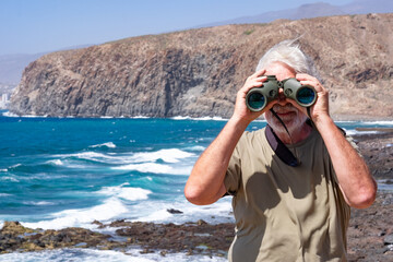 Front view of senior man holding binoculars looking smiling at camera. Senior white haired pensioner enjoying retirement at sea. Cliffs and mountain in background