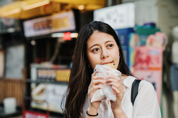 hungry charming asian japanese girl trying taiwanese local street food in plastic bag and looking aside under sunshine. beautiful lady tourist enjoy traditional snack outdoor market in summer time