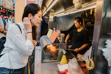 young asian korean girl tourist travel in taichung taiwan outdoor market. lady buying street food on vendor stall and curiously watching how seller cooks. beautiful female looking at snack stand