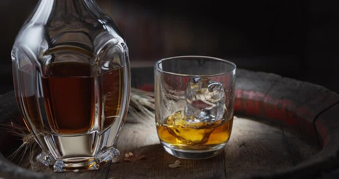 An ice cube slowly falls into a glass of whiskey on top of a vintage whiskey barrel. A decanter with a drink stands nearby, a dark brown background. 