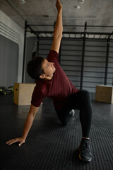 Fototapeta na wymiar Man warming up on floor at gym before crossfit training standing on one knee and straight hand, stretching leg and putting left hand up, twisting back, dressed in black tights and red fitness T-shirt