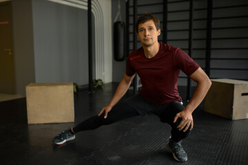 Man in sportswear and sneakers stretching legs sitting in squat position, trying to balance holding hands on knees, looking in front of him, breathing deeply, doing daily workout to keep fit