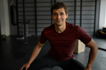 Obraz premium Close-up of cute smiling man of 30s in stylish fitness wear doing squats on leg day at gym, balancing, looking happy and satisfied with exercises, posing against sport equipment of gym