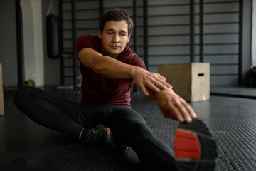 Horizontal picture of man trainig at gym, sitting on floor in sneakers and tights, wearing red T-shirt, reaching hand to toe, stretching left leg, keeping right foot near inner side of hip