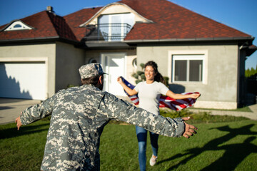 Soldier in uniform coming home and his lovely wife with American flag running into his arms celebrating reunion.