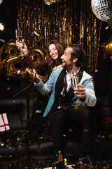cheerful couple with champagne celebrating new year on sofa near shiny decoration on black