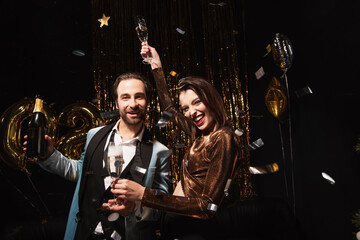 joyful couple with bottle and glasses of champagne looking at camera near christmas decoration on...