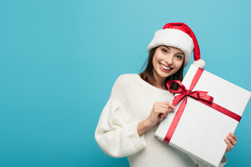 joyful woman in santa hat looking at camera while holding present isolated on blue
