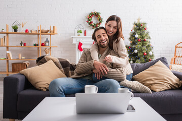 happy woman hugging husband and pointing at laptop on sofa in living room