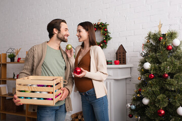 cheerful man with wooden box looking at wife holding baubles near christmas tree