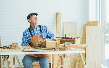 Senior old aging beard kind Caucasian man wearing check shirt, apron, making DIY wooden furniture, smiling with happiness, looking away at window, sitting alone at home. Retirement, Hobby Concept.
