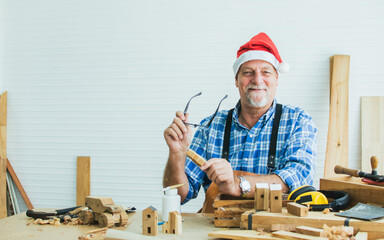 Portrait senior old aging kind Caucasian man or carpenter wearing santa hat, making DIY wooden furniture, holding eyeglasses, smiling with happiness, sitting alone at home. Retirement, Hobby Concept.