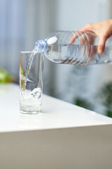 Closeup image of a female hand holding drinking water bottle and pouring water into glass on table.