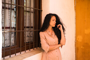 Fototapeta premium beautiful woman with dark curly hair leaning against the wall and the window grille of a typical Mediterranean style house. The woman is serious and sad. Concept expressions.