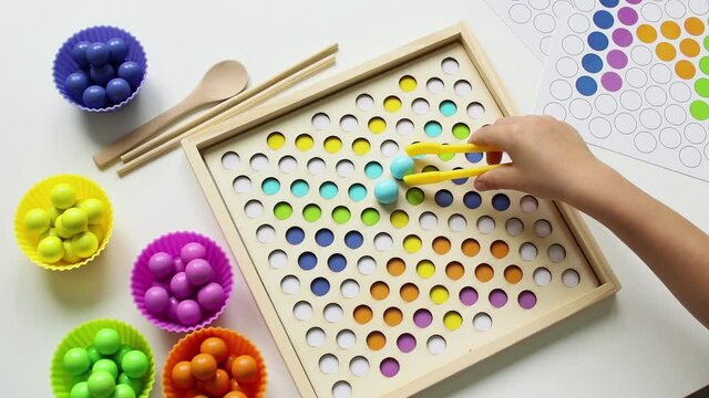 The child lays out colorful balls with tweezers. Wooden toy. Study of colors. Montessori concept, early brain exercise. Early education, fine motoric skills. Kids leisure. Preschool education.