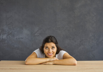 Cheerful lady imagining her future and smiling. Portrait of happy young woman sitting at wooden table, resting chin on hands, looking up, dreaming, thinking about something pleasant