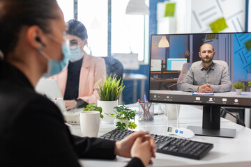 Businesswoman with medical face mask against covid19 discussing financial strategy with remote manager during online videocall meeting conference in startup office. Teleconference on computer screen
