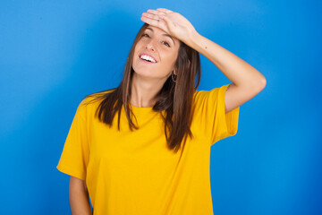 Fototapeta na wymiar Young european brunette woman wearing yellow T-shirt on blue backgroundwiping forehead with hand making phew gesture, expressing relief feels happy that he prevented huge disaster. It was close enough