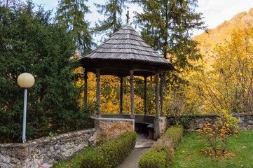 Wooden gazebo for relaxing in a city park surrounded by trees with green and yellow leaves. Rustic pavilion with a cross above in the autumn park