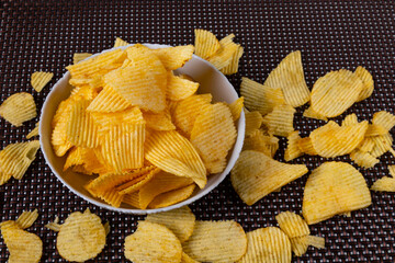 Potato chips are in a white cup on a brown tablecloth. Chips are also scattered nearby.