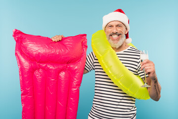 Obraz premium cheerful middle aged man in santa hat and swim ring holding pink inflatable mattress and glass of champagne isolated on blue