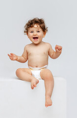 Vertical portrait of little cute toddler boy, baby in diaper joyfully sitting and laughing isolated over white studio background. Sweet childhood - obrazy, fototapety, plakaty