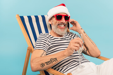 cheerful mature man in santa hat holding glass of champagne while talking on smartphone and sitting on deck chair isolated on blue