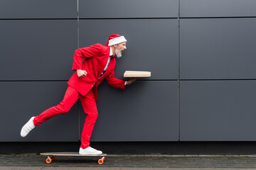 side view of mature man in santa hat holding pizza box and riding on skateboard near grey wall on urban street
