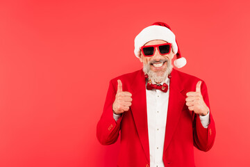 happy mature man in santa hat and sunglasses with christmas balls on beard showing thumbs up isolated on red