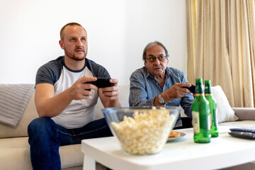 Father and his son spending their free time playing video games and enjoying each others company. Overjoyed senior father sit relax on couch with son playing computer game with joysticks.