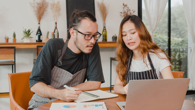 Stressed Asian Small Business Owners Couple Using Laptop Discussing Project And Finance With Documents Of Shop For The Month At Cafe. Woman And Man Doing Paperwork Together, Paying Taxes Online.