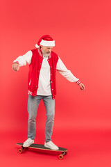 Fototapeta na wymiar full length of middle aged man in santa hat riding skateboard on red