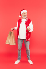 full length of bearded middle aged man in santa hat holding shopping bags while using smartphone on red