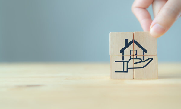 Home loan, mortgage and buy a real estate concept. Hand hold wood cube on the table with icon of hands holding house model in grey background and copy space.Banner for home buying, loan and insurance.