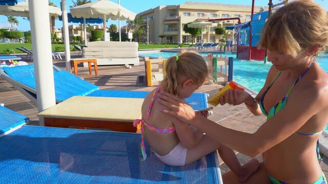 The child is sitting on a sun lounger near the pool. Mom puts sunscreen on the baby's back. 