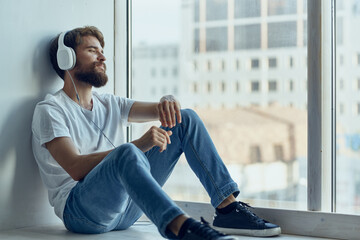 man in white t-shirt sitting near the window in headphones listening to music
