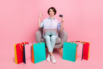 Full length body size photo woman sitting with packages showing credit card okay sign with laptop isolated pastel pink color background