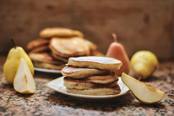 Pear pancakes sprinkled with powdered sugar next to ripe pears. A bunch of pancakes in the background.