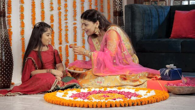 Smiling young mother feeding sweets to her cute daughter during festivities. Traditional Rangoli and a colorful gift box kept together - Diwali decorations  celebration time  a Hindu festival  fami...