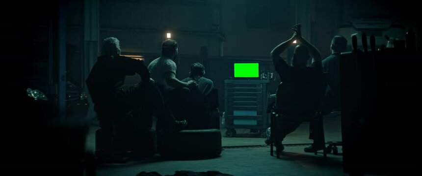 WIDE Car garage workshop workers in uniforms watching portable TV in the evening, sport event, unhappy with a result. Green screen, chroma key. Shot with 2x anamorphic lens