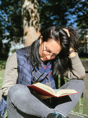 Young woman is reading red book in a park. Young candid millennial woman in casual autumnal clothes siting on a wooden bench in park and reading red book. Concept of back to school or university 