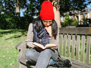 Fototapeta na wymiar Young candid woman in casual autumnal clothes siting on a wooden bench in park and reading red book. Early autumn. Wearing red hat and beige woollen sweater. Happy brunette, confident, generation z