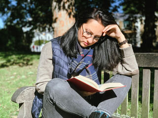 Young woman is reading red book in a park. Young candid millennial woman in casual autumnal clothes siting on a wooden bench in park and reading red book. Concept of back to school or university 