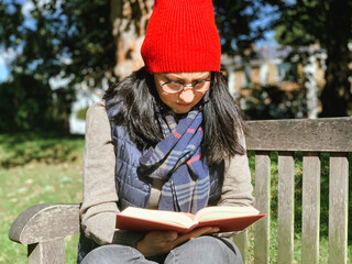 Young candid woman in casual autumnal clothes siting on a wooden bench in park and reading red book. Early autumn. Wearing red hat and beige woollen sweater. Happy brunette, confident, generation z