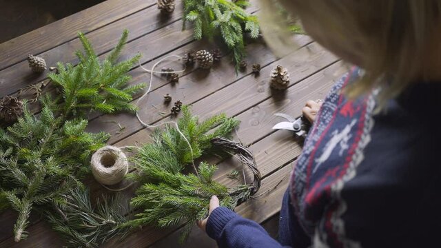 Aged wooden table in authentic florist workshop. Backside view female arrange pine cone and attache evergreen fir branches preparing handmade festive decoration. DIY and handcraft concept
