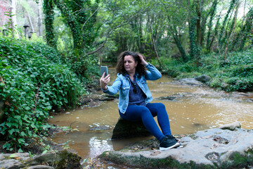 Woman sitting on a rock next to a stream running through a gallery forest, takes a selfie with her smartphone, while holding her hair in her hand.