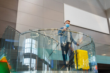 Young caucasian man with suitcase waiting at airport in waiting room for his departure. Man in protective face mask with luggage In airport lounge balcony with outdoor advertising mockup
